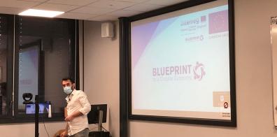 Man in facemask standing in front of screen displaying BLUEPRINT Project powerpoint presentation